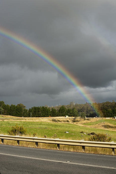 View from a tarred road of a colorful rainbow in a country field with stormy grey skies above