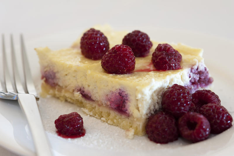 Slice of baked ricotta cheesecake topped with fresh ripe raspberries with a serving of raspberry compote on the side