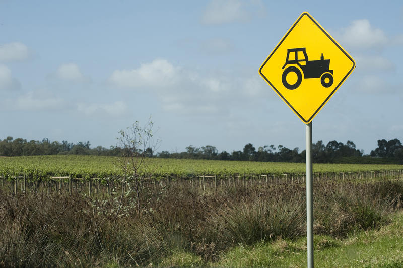 Yellow traffic warning sign for agricultural vehicles and equipment showing the silhouette of a tractor at the roadside alongside an agricultural field