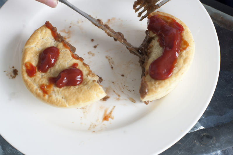 High angle view of a person eating a savory meat pie for lunch with gravy on the pastry crust cutting it with a knife and fork