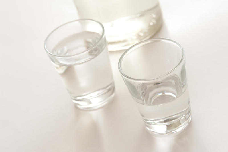 White spirits in a shot glass, one half finished and one full with the bottom of a bottle of alcohol visible behind