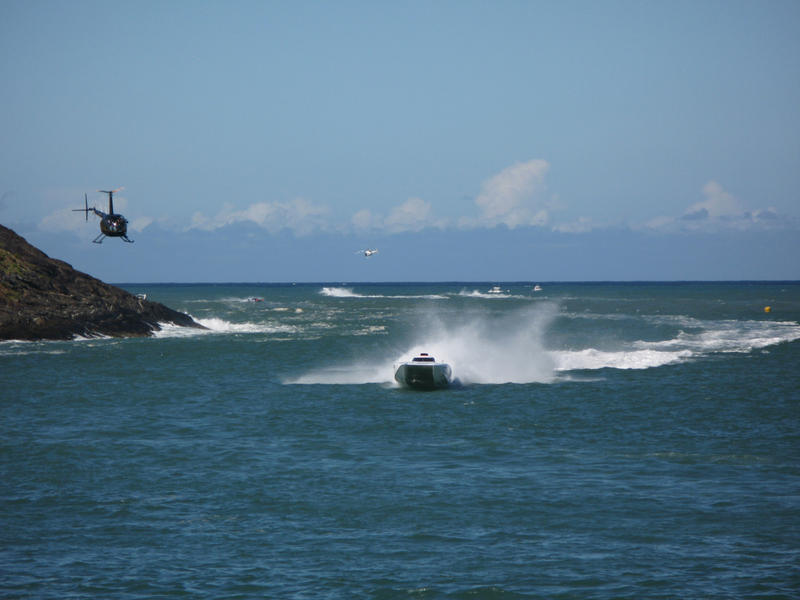 Offshore power boat racing with a speedboat speeding towards the camera along the coastline with a helicopter hovering above