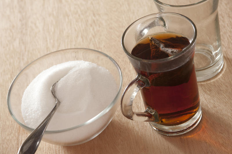 Sugary sweet hot black tea in a glass mug with a bowl full of white sugar and a teaspoon alongside on a wooden table, high angle view