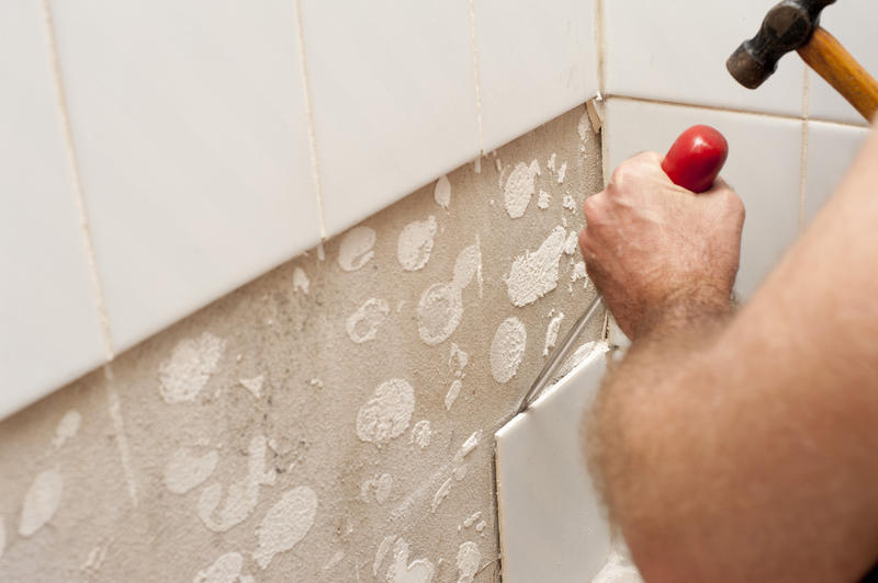 Man removing old white ceramic wall tiles with a screwdriver and hammer, close up of his hands