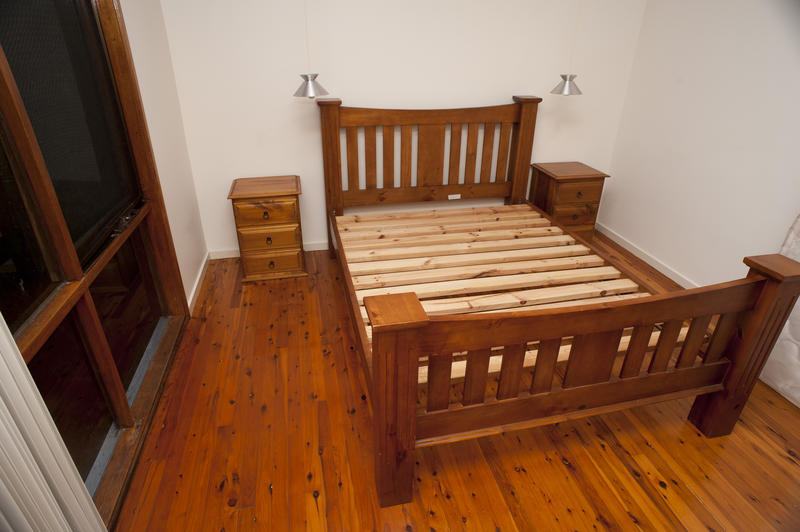 Classis wooden bed frame with a slatted headboard and footboard standing in a small bedroom on a bare hardwood floor alongside a window