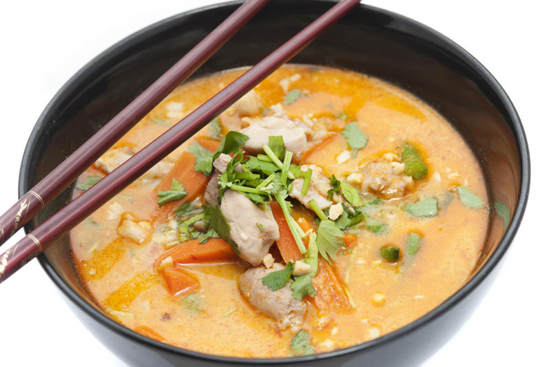 Close up view spicy laksa soup garnished with green onions and herbs in little black bowl with deluxe brown chopsticks