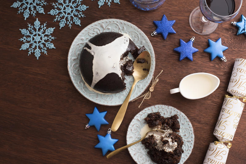 Serving Christmas pudding with brandy cream on a festive blue themed table with stars and snowflakes viewed from overhead with a served plate to the side