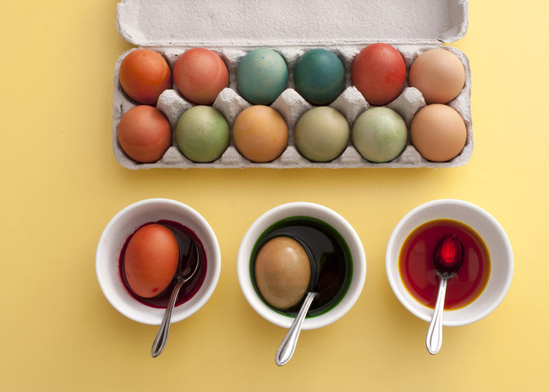 Preparing dyed colored eggs for Easter steeping them in three bowls of dye with a full cardboard carton of finished eggs alongside, overhead view