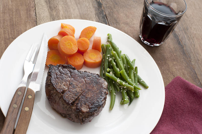 Hearty and healthy dinner of well done ribeye steak besides cooked carrot pieces and seasoned green beans by glass with refreshing beverage