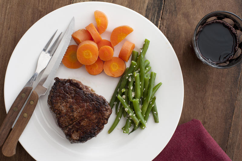 Medallion of grilled fillet steak with fresh vegetables, diced carrots and seasoned green beans, served on a plate with a glass of red wine on the side, overhead view