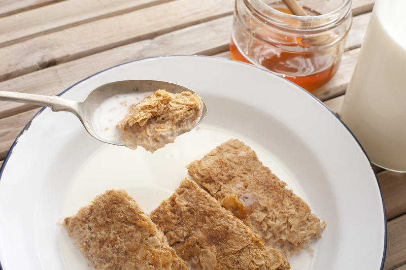 Bowl filled with shredded wheat biscuits and milk besides jar of honey on a wooden table