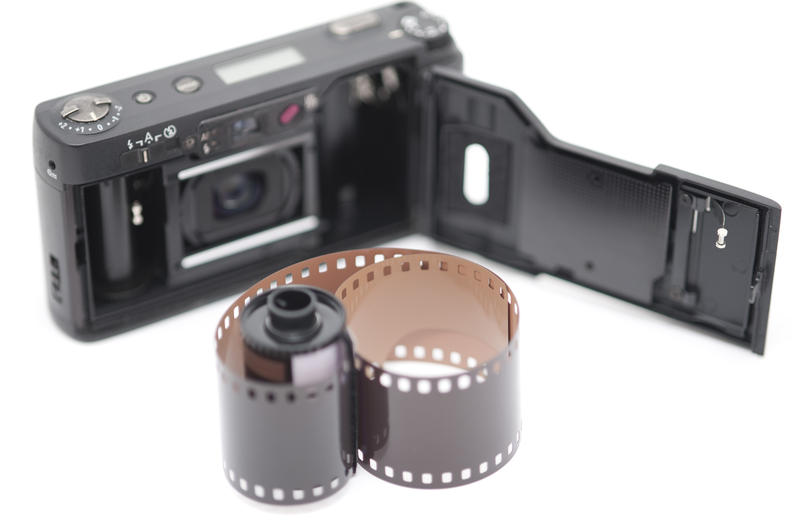 Single exposed roll of film next to compact 35 millimeter pocket camera with open back cover over white background in selective focus