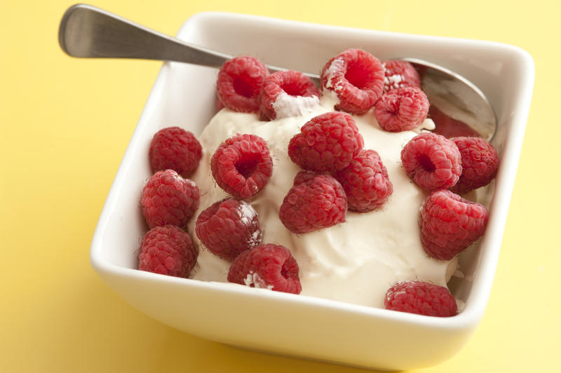 Fresh seasonal raspberries on plain yoghurt served in a square white bowl for a healthy breakfast or dessert on a yellow background