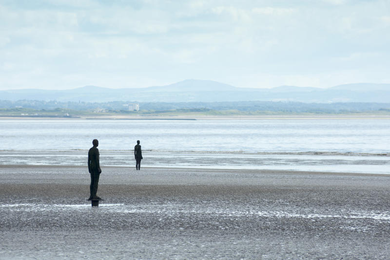 Another Place by Antony Gormley, an installation of cast iron figures on Crosby Beach, UK standing facing out over the ocean in a travel concept, with copy space