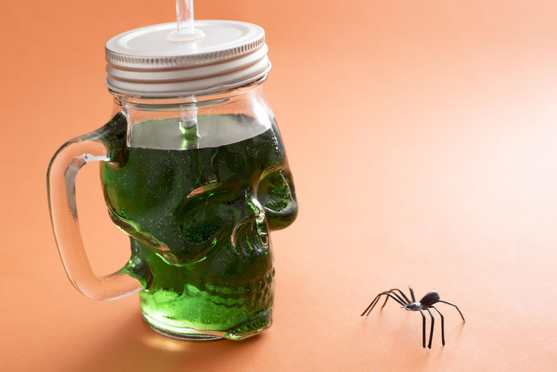 Spooky green Halloween drink in a skull shaped Mason jar with lid facing a creepy spider on an orange background with copy space