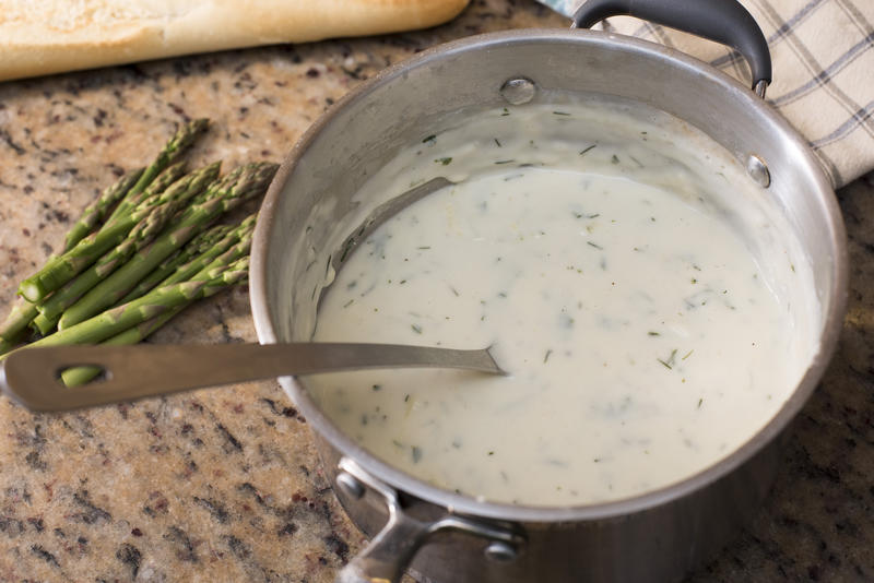 Stainless steel pot with herbed white sauce and spoon beside a small bunch of asparagus