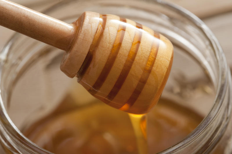 Close up on a wooden honey dipper coated in honey held above an open honey jar