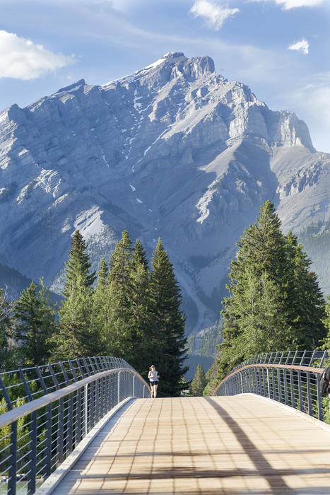 <p>A female jogger runs over a wooden bridge in Banff, Canada with the Rocky Mountains in the background</p>
