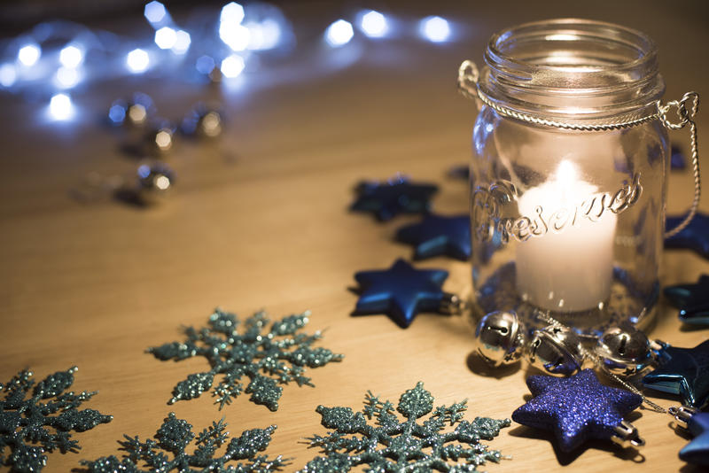 Burning candle in a glass jar surrounded by Christmas ornaments on a wooden table with copy space and background bokeh of sparkling party lights