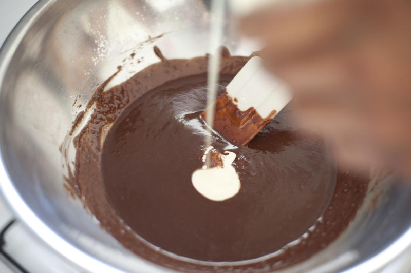Baker making chocolate icing in a stainless steel mixing bowl pouring the icing sugar into the melted chocolate while stirring with a spatula