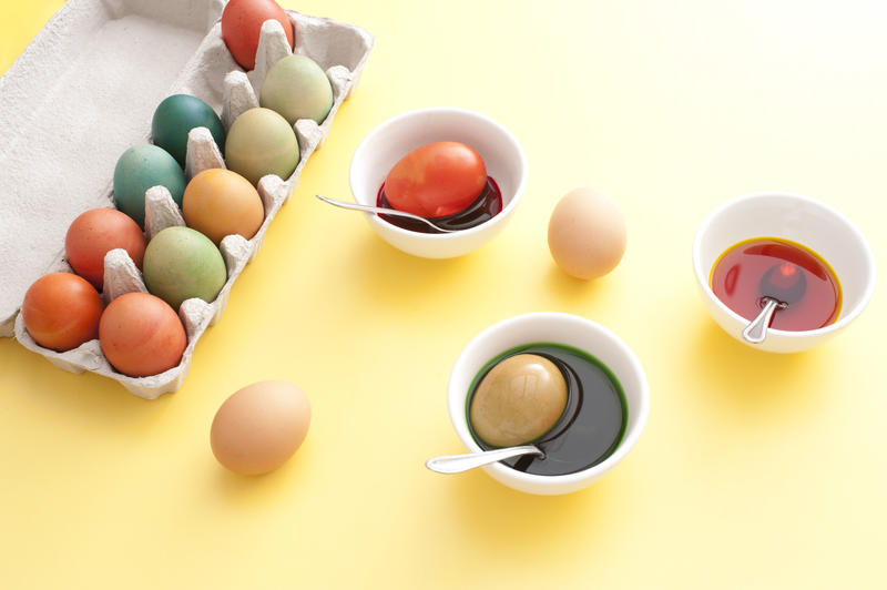 Process of dyeing Easter eggs in white bowls with liquid paint, spoons and box of colored eggs on yellow table surface background