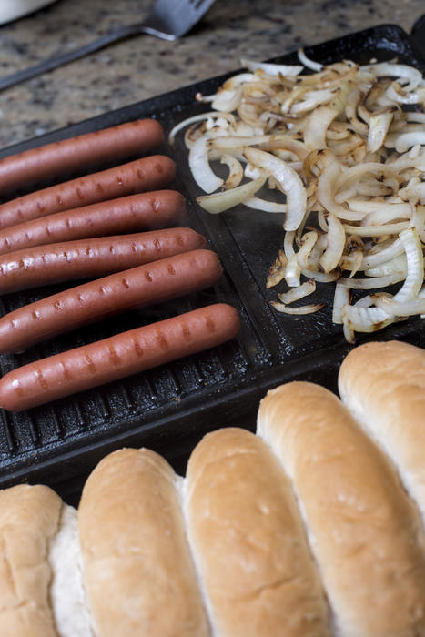 Scene of various hot dogs being prepared on grill with onions and uncut bread buns