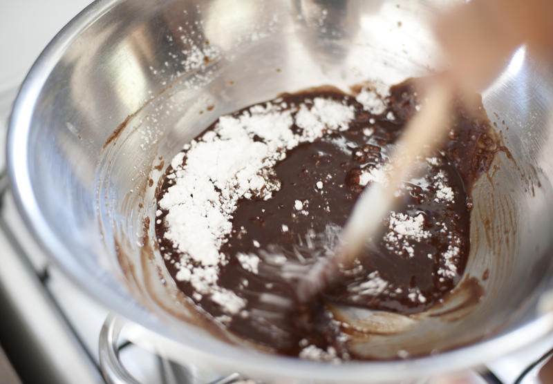 Person making chocolate icing for a cake with a view into the mixing bowl of melted chocolate and sprinkled icing sugar being blended with a wooden spoon with motion blur