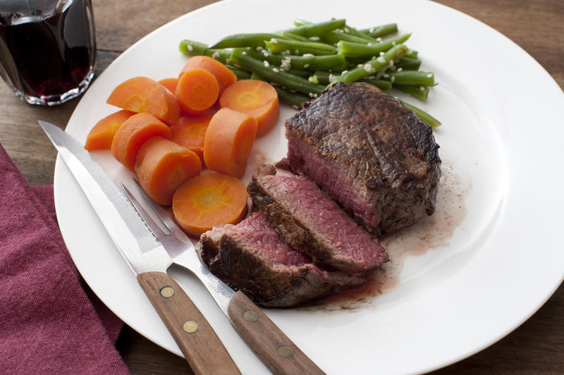 Thick juicy medallion of medium rare fillet steak with fresh carrots and green string beans served with a glass of red wine and utensils