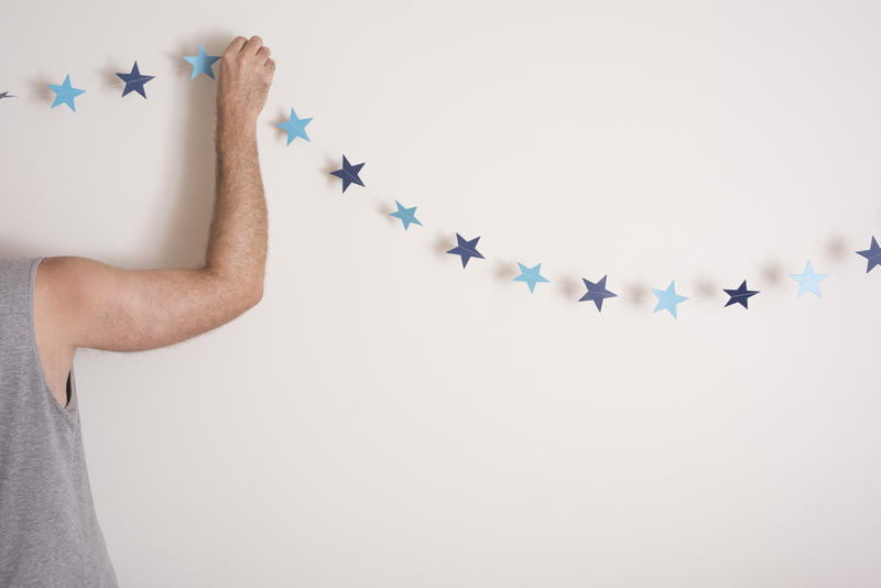 Man decorating his house for a minimalist Christmas hanging a simple string of blue paper stars or bunting on his wall, with copy space