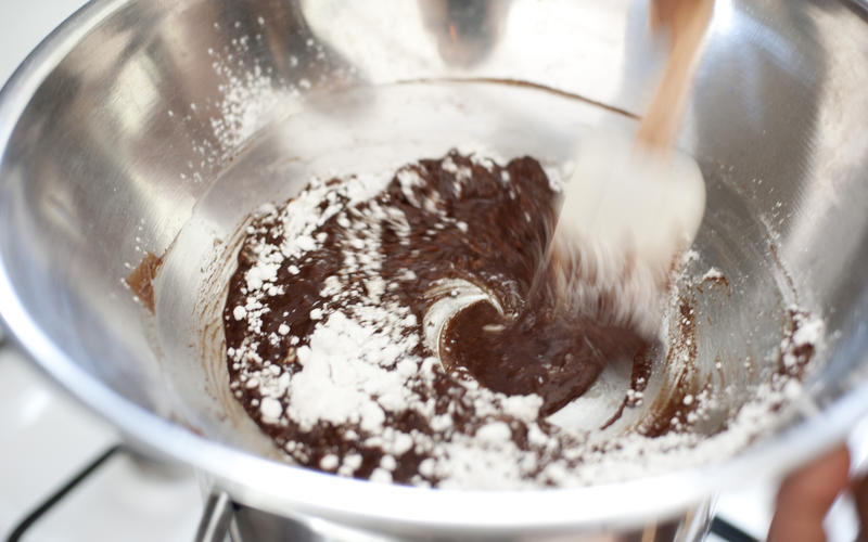 Mixing a bowl of chocolate icing pouring the icing sugar onto melted chocolate and blending with a wooden spoon