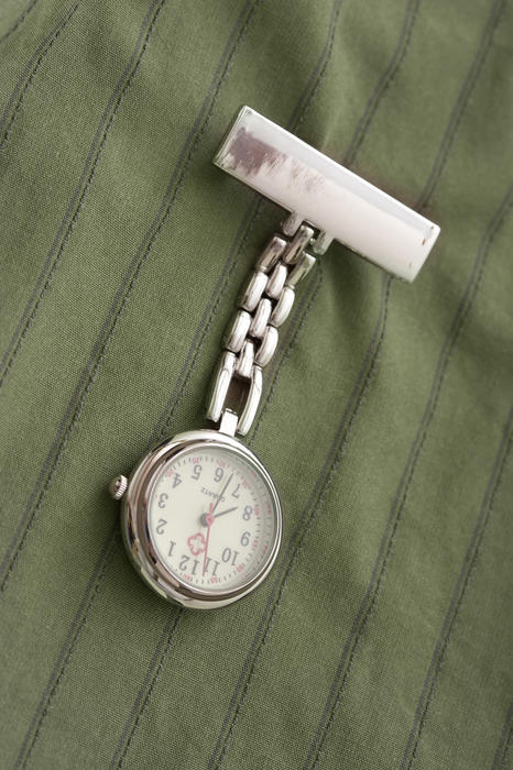 Stainless steel nurses watch with a blank name label pinned to a green striped uniform hianging down on its chain diagonally through the frame
