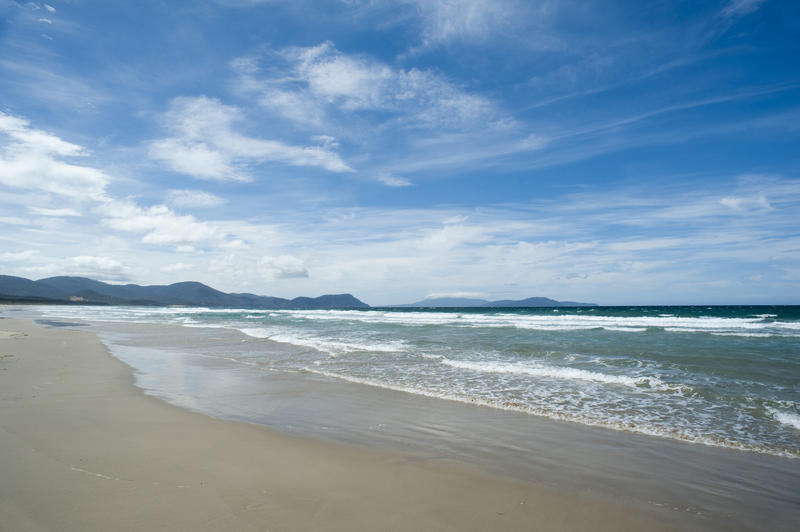 Long sandy tropical beach with gentle waves breaking on the shore and a distant headland under a cloudy blue sky