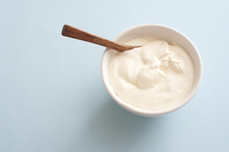 Small white round bowlk of plain yogurt with wooden spoon against a light colored background