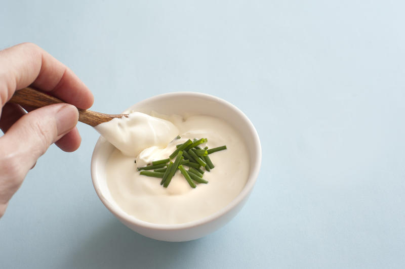 Man dipping a small spoon into a white ceramic bowl of sour cream garnished with fresh chopped chives over a grey background with copy space