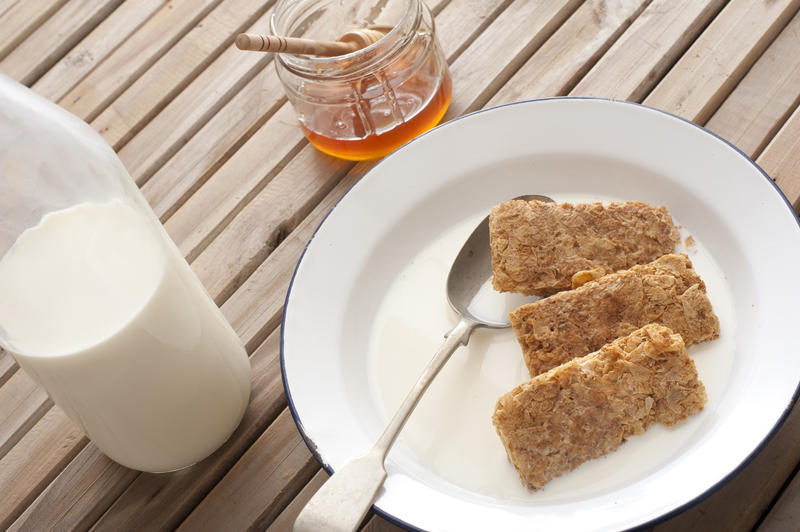 Three wheat biscuits of breakfast cereal in milk with spoon besides glass bottle and jar of honey