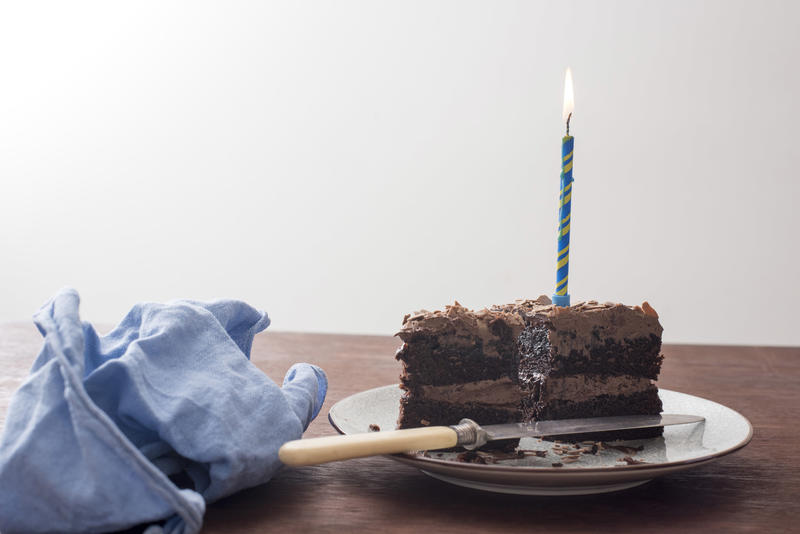 Chocolate birthday cake with burning blue candle on a plate on a table with matching blue napkin viewed low angle