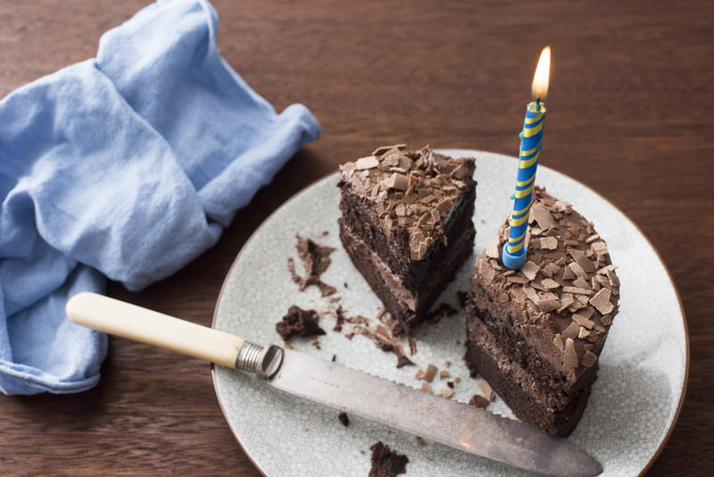 Cut iced chocolate birthday cake with single burning blue candle and matching napkin on a wooden table