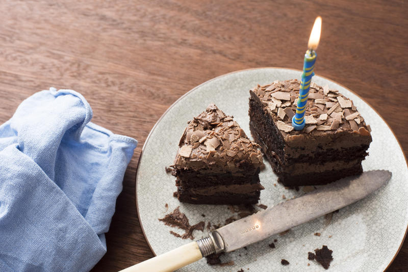 Remaining slices of a chocolate birthday cake with burning blue candle on a white plate with knife on a wooden table