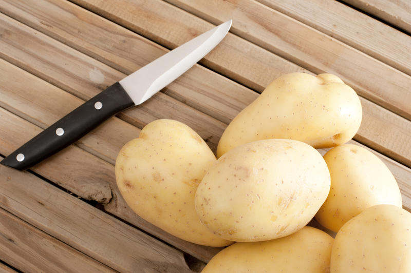 A close up of white potatoes and a sharp knife on a timber slatted bench.