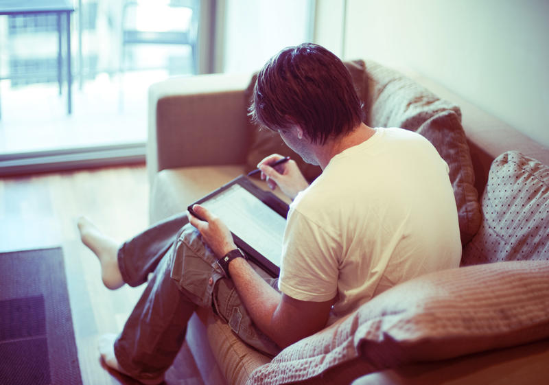Young businessman working from home during Covid-19 seated on a sofa in the living room using a tablet pc viewed from the rear